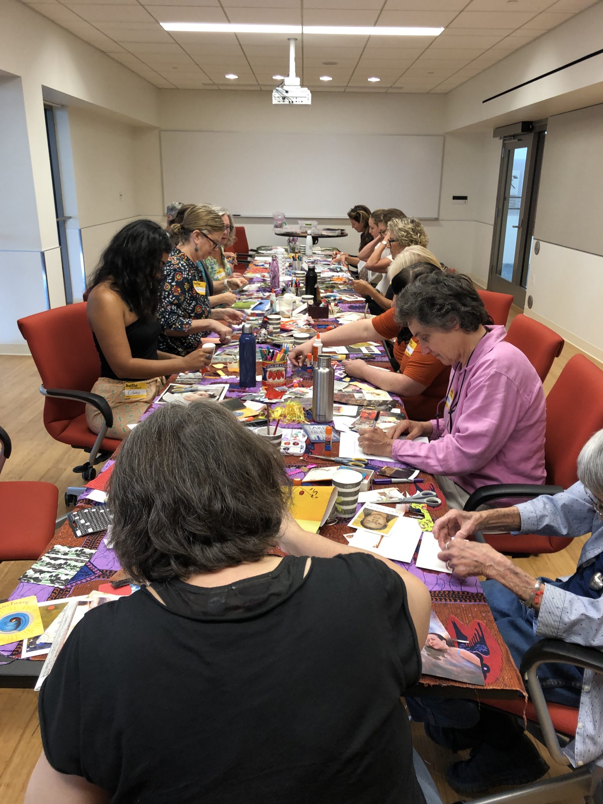 A group of women seated at a long conference table, making postcards with collage materials.